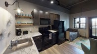 This single-floor studio unit showcases an industrial-chic aesthetic that blends raw architectural elements with modern finishes. The open-plan layout seamlessly connects the kitchen, living, and sleeping areas into one cohesive living space, making efficient use of every square foot.

The kitchen is outfitted with dark espresso cabinetry, black appliances including a range, over-the-range microwave, and refrigerator, and features striking marble-look countertops. Open wooden shelving provides additional storage and display space, adding a warm, rustic contrast to the darker cabinetry. Light wood-tone flooring runs throughout, enhancing the sense of openness.

The living and sleeping area benefits from soaring ceilings with exposed ductwork and industrial-style track lighting, lending the space a loft-like character. A set of multi-pane French doors floods the room with natural light and offers a direct connection to the exterior. The tall ceilings further amplify the airy, urban loft ambiance.

The bathroom is clean and well-appointed, featuring a dark vanity with a white countertop, wood-look flooring, and a shower-tub combination enclosed by a colorful curtain. The neutral wall tones keep the space feeling bright and functional.

Overall, this studio presents a thoughtfully designed urban living environment with strong industrial character and practical, modern amenities throughout.