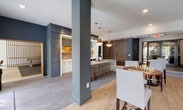 Located at 101 E Romana St in downtown Pensacola, Florida, this single-floor amenity space is a well-appointed community lounge designed for both relaxation and social engagement. The interior showcases a contemporary aesthetic, blending textured accent walls — including a striking gray shiplap feature wall — with warm wood-look flooring, patterned carpet, and a refined neutral color palette accented by deep blues and natural wood tones.

The lounge area serves as the heart of the space, furnished with a plush sectional sofa, vibrant blue wingback chairs, and a sculptural live-edge root console table that adds an organic, artistic touch. A wall-mounted TV anchors the seating area, while large windows invite natural light and offer outdoor views of the surrounding grounds.

The full kitchen is outfitted with white cabinetry, a generous marble-topped island with bar seating, and pendant lighting with gold brass fixtures — ideal for catering or community gatherings. Slatted wood partitions elegantly define zones throughout the open floor plan.

A dedicated game room provides a pool table, shuffleboard table, wall-mounted TV, and high-top seating, all framed by warm wood slat screens. Additional seating nooks with round tables offer space for board games and casual conversation, completing a versatile and inviting amenity experience.