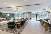 Located at 4720 Nations Crossing Rd in Charlotte, NC, this generously sized single-floor amenity space offers a sophisticated and thoughtfully designed communal environment. The open-plan layout is anchored by large floor-to-ceiling windows and glass doors that flood the interior with natural light and provide views of the surrounding outdoor areas. Light-toned large-format tile flooring flows throughout, unifying the various distinct zones within the space.

The main lounge area features plush sectional seating, accent armchairs in rich jewel tones, and designer chandeliers with globe pendants that add warmth and visual interest overhead. Wood-clad columns punctuate the open floor plan, adding an organic texture that complements the contemporary aesthetic.

A cozy lounge nook is defined by a striking freestanding suspended fireplace at its center, surrounded by upholstered seating and intimate booth-style banquettes beneath a wood-paneled ceiling. An adjacent wall showcases a dramatic honeycomb moss installation set against exposed brick, providing a vibrant biophilic focal point alongside curated artwork.

A coffee and beverage bar station with white cabinetry and a decorative herringbone tile backsplash serves as a functional amenity hub. Bold geometric partition screens and living plant walls further divide the space into inviting zones, creating a dynamic yet cohesive environment suited for both social gatherings and quiet retreat.