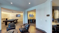 The game room is a well-appointed, multi-zone entertainment space designed for both casual socializing and competitive play. The main area features hardwood-style flooring and is anchored by a full-size pool table, complemented by plush leather lounge chairs that invite relaxed gathering. A wall-mounted flat-screen television is positioned for easy viewing from the seating area, making the space suitable for watching sports or gaming.

A textured stone accent wall runs along one side, adding warmth and visual depth to the otherwise clean, contemporary aesthetic defined by white walls and recessed lighting. The smooth transition between the main lounge area and an adjacent private room is framed by a wide archway. This secondary space features a dining-height table surrounded by upholstered chairs, its own wall-mounted display, and a pendant light fixture overhead — ideal for group gaming, card nights, or casual dining. The stone wall treatment continues into this room, creating a cohesive design language throughout. Together, the two zones offer a versatile and stylish environment for a variety of recreational activities.