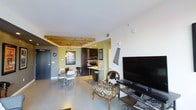 This single-floor space offers a thoughtfully designed open-concept layout that seamlessly connects the living, dining, and kitchen areas. The living area serves as a central gathering point, featuring an eclectic mix of furnishings including a distinctively upholstered accent chair, a large flat-screen TV, and decorative wall art that adds personality to the space. Polished concrete flooring runs throughout, lending the interior a contemporary, industrial feel while remaining easy to maintain.

A standout architectural detail is the exposed natural wood beam ceiling treatment that frames the transition between the living and dining areas, adding warmth and visual interest to the otherwise clean, white ceiling. A pendant light fixture hangs above the dining area, which is furnished with a sleek table and modern white chairs, creating an intimate yet functional space for meals.

The kitchen is finished in dark cabinetry with stainless steel appliances, including a built-in microwave and range, and features a breakfast bar with counter-height seating — ideal for casual dining. Recessed lighting illuminates the kitchen efficiently. The overall ambiance of the space balances modern industrial elements with warm, eclectic touches, resulting in a dynamic and inviting environment.