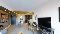 This single-floor space offers a thoughtfully designed open-concept layout that seamlessly connects the living, dining, and kitchen areas. The living area serves as a central gathering point, featuring an eclectic mix of furnishings including a distinctively upholstered accent chair, a large flat-screen TV, and decorative wall art that adds personality to the space. Polished concrete flooring runs throughout, lending the interior a contemporary, industrial feel while remaining easy to maintain.

A standout architectural detail is the exposed natural wood beam ceiling treatment that frames the transition between the living and dining areas, adding warmth and visual interest to the otherwise clean, white ceiling. A pendant light fixture hangs above the dining area, which is furnished with a sleek table and modern white chairs, creating an intimate yet functional space for meals.

The kitchen is finished in dark cabinetry with stainless steel appliances, including a built-in microwave and range, and features a breakfast bar with counter-height seating — ideal for casual dining. Recessed lighting illuminates the kitchen efficiently. The overall ambiance of the space balances modern industrial elements with warm, eclectic touches, resulting in a dynamic and inviting environment.