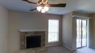 This single-floor home offers a comfortable and functional layout designed for everyday living. The living area serves as the heart of the space, highlighted by a classic brick fireplace with a clean white mantle that adds a traditional, warm character to the room. Soft, neutral-toned walls complement the carpeted flooring, creating an inviting atmosphere throughout. A ceiling fan with an integrated light fixture is mounted overhead, providing both ambient lighting and year-round climate comfort. Natural light filters into the living area through a window dressed with horizontal blinds, offering privacy while still brightening the interior. The textured ceiling adds a subtle architectural detail that complements the overall aesthetic. The space presents a clean, move-in ready environment with a straightforward, practical design. The brick surround of the fireplace stands as a standout focal point, grounding the living area with a sense of warmth and homeliness. Overall, the space combines classic design elements with comfortable, livable features suited for a relaxed lifestyle.