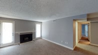This single-floor space presents a straightforward and functional layout centered around a generously sized living area. The room is finished with wall-to-wall beige carpeting that adds warmth underfoot, complemented by neutral gray walls and crisp white baseboards that give the interior a clean, understated aesthetic. A textured popcorn ceiling adds a classic touch overhead, while a ceiling-mounted HVAC vent ensures year-round climate comfort.

A built-in fireplace with a simple white mantel serves as a natural focal point along one wall, providing both visual interest and a cozy element to the living space. Adjacent to the fireplace, a rear-facing door with a window panel allows natural light to filter into the room, brightening the interior and offering potential access to an outdoor area.

The open wall space provides flexibility for a variety of furniture arrangements, making the room adaptable to different living needs. A doorway on the far side of the room suggests additional spaces beyond, contributing to the overall flow of the single-level floor plan. The space offers a neutral, move-in-ready canvas suitable for a range of interior styles.