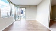Located at 809 S Washington St in Seattle's Pioneer Square neighborhood, this 490-square-foot studio unit offers a thoughtfully designed single-floor layout comprising a combined kitchen and living area, a separate living room, and one bathroom. The living area features light wood-tone laminate flooring throughout, neutral warm-gray walls, and large corner floor-to-ceiling windows with a sliding glass door that opens to views of the Seattle skyline and waterfront. A secondary window also frames partial water and port views, filling the space with natural light. Track lighting and a pendant light fixture provide additional illumination. The kitchen is outfitted with contemporary flat-panel cabinetry in a dark wood-grain finish, light quartz countertops, a glass tile backsplash, under-cabinet lighting, and a full suite of stainless steel appliances including a refrigerator, electric range, over-the-range microwave, and dishwasher. The bathroom features a glass-enclosed walk-in shower with white subway tile surround, brushed nickel fixtures, an LED-lit vanity mirror, and an in-unit stacked washer and dryer. A hallway connects the kitchen to the entry door, maintaining a clean, efficient flow throughout the unit.