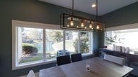This single-floor residence combines modern design elements with a warm, inviting ambiance throughout its thoughtfully arranged layout. The dining area serves as a standout feature of the home, anchored by a striking industrial-style rectangular pendant light with exposed Edison bulbs encased in a metal and glass frame, casting a rich, ambient glow over the dining table. Deep green-toned walls add depth and sophistication to the space, complementing the natural wood tones of the dining table and the sleek black upholstered chairs surrounding it. Large picture windows frame the dining area, flooding the room with abundant natural light and offering picturesque views of a tranquil pond and lush greenery beyond the property. This seamless connection between the interior and the outdoor landscape creates a peaceful, nature-inspired atmosphere that enhances the overall character of the home. The single-floor design ensures ease of movement and accessibility throughout, while the cohesive use of materials and finishes gives the space a curated, polished feel. Whether used for everyday meals or entertaining guests, the dining area exemplifies the home's blend of functional design and aesthetic appeal.