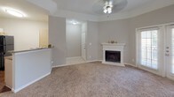 This single-floor residence offers a well-organized layout with distinct living, sleeping, and utility areas. The living room serves as the central gathering space, featuring a classic white-mantled fireplace, soft wall-to-wall carpeting, a ceiling fan with light fixture, and a tray ceiling detail that adds architectural interest. An open pass-through connects the living area to the kitchen, promoting an open flow between spaces. The kitchen is equipped with white cabinetry, granite-look countertops, a double stainless steel sink, a mosaic tile backsplash, and hardwood-style flooring, lending a functional yet polished look. A dedicated laundry area is accessible off a hallway, featuring washer and dryer hookups alongside a walk-in closet with wire shelving, offering practical built-in storage. The bedroom is generously proportioned with plush carpeting, a ceiling fan, and a window fitted with horizontal blinds that allow natural light while maintaining privacy, with views of mature outdoor greenery. Additional rooms feature crisp white walls, crown molding, and neutral tones throughout, creating a cohesive and move-in ready environment. The overall ambiance is clean, neutral, and versatile, suited to a variety of living arrangements.