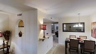 Located in Euless, TX, this single-story space offers a thoughtfully arranged open layout that promotes a natural flow between living areas. A defining architectural feature is the half-wall pass-through counter, which connects the kitchen or serving area to the adjacent spaces, making it ideal for entertaining or everyday convenience. The counter is accented by decorative corbel brackets, adding a subtle traditional touch to the otherwise neutral interior.

The dining area is positioned alongside a large, dark-framed wall mirror that enhances the sense of depth and reflects ambient light throughout the space. A dining table with upholstered chairs is visible, providing a functional yet comfortable setting for meals. Neutral-toned carpeting runs throughout the living areas, contributing to a warm and cohesive aesthetic.

The walls are finished in a clean, off-white tone that keeps the space feeling bright and airy. Recessed ceiling vents and standard lighting fixtures are integrated throughout. A hallway leads further into the unit, suggesting additional rooms beyond the main living and dining areas. Overall, the space presents a practical and welcoming single-floor layout suitable for everyday living.
