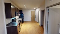This single-floor unit is designed with a practical and modern layout, offering a comfortable living experience through its thoughtfully arranged spaces. The kitchen serves as a standout feature, showcasing rich dark espresso cabinetry extending from floor to ceiling, providing generous storage. Complementing the cabinetry are dark granite-style countertops, a stainless steel double sink, and a full suite of black appliances, including an electric range, built-in microwave, dishwasher, and refrigerator. Track lighting overhead illuminates the kitchen efficiently. Light-toned hardwood-style flooring runs through the kitchen and open living area, creating a bright and cohesive feel throughout the main space. The entryway opens directly into this combined area, with a keypad-secured front door and a utility panel nearby. The bedroom offers a quieter retreat, finished with plush neutral carpeting, a closet, and understated gray-toned walls. The bathroom is clean and functional, featuring dark tile flooring, a tub-shower combination, a white vanity with a single sink, dark cabinetry below, and a well-lit mirror above. The overall aesthetic is contemporary and consistent, with a neutral palette carried throughout.