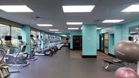 This fitness center offers a spacious, single-level layout designed to accommodate a full range of workout activities. The cardio zone is prominently featured along one side of the room, with a row of treadmills and elliptical machines lined up facing large windows that invite natural light and provide views of outdoor greenery, creating an energizing atmosphere for exercise.

The interior design incorporates a contemporary color scheme, with teal accent walls contrasting against dark baseboards and charcoal-toned flooring. The drop ceiling is outfitted with recessed panel lighting that evenly illuminates the entire space, ensuring excellent visibility throughout. Rubber flooring covers the workout areas, providing durability and support underfoot.

A structural column with matching teal finishes divides sections of the space, adding visual interest while maintaining an open feel. Dark shelving units offer storage options, and a separate room with frosted glass doors can be seen at the far end, likely providing access to private or auxiliary spaces. The overall ambiance is clean, modern, and functional, making it a practical environment for fitness activities.
