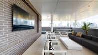 This rooftop terrace lounge offers an elevated urban retreat with sweeping panoramic views of a vibrant city skyline. The space is designed for comfort and socializing, featuring a generous open-air layout partially covered by a sleek paneled ceiling supported by clean white columns, seamlessly blending indoor and outdoor living.

The main lounge area is furnished with a mix of contemporary white-framed seating with light gray cushions and a large, low-profile sectional sofa in charcoal gray accented with gold and striped throw pillows. Multiple seating groupings are arranged to take full advantage of the unobstructed skyline views, while lush tropical potted plants add a refreshing natural element throughout.

A standout feature is the striking dark brick accent wall, which anchors one side of the terrace and hosts a wall-mounted flat-screen television, creating an ideal setting for both relaxed lounging and social gatherings. A built-in spa or hot tub is also visible near the brick wall, adding a luxurious amenity to the space.

Access to the terrace is through double glass doors framed by the same brick exterior, lending a cohesive architectural character. Just inside, a sleek dark reception or service counter sits beneath a bright window overlooking a lush green landscape, providing a functional transition space. Large-format light tile flooring runs throughout, unifying the entire level with a clean, modern aesthetic.