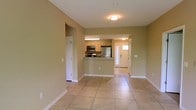This single-story space features an open-concept design that seamlessly connects the living area to the kitchen, creating a cohesive and functional floor plan. Light beige ceramic tile flooring extends throughout, contributing to a clean, consistent aesthetic. The walls are painted in a soft, warm green-beige tone, lending the interior a neutral and welcoming ambiance.

The kitchen is positioned at the center of the space and is defined by a raised breakfast bar that serves as a natural divider between the cooking and living areas. It is equipped with light wood cabinetry, a stainless steel refrigerator, a microwave, and a range, providing practical functionality. Under-cabinet lighting illuminates the counter workspace effectively.

Flush-mounted ceiling light fixtures provide overhead illumination in both the living and kitchen areas. A doorway visible toward the rear of the space leads to an entry area with a windowed door that allows natural light to filter in. Additional doorways suggest access to further rooms or storage areas. White baseboards and trim throughout provide a crisp contrast to the neutral wall color, completing the tidy and straightforward interior design.