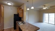 This single-floor space showcases a thoughtfully designed open-concept layout that connects the kitchen and living area in a seamless, functional arrangement. The kitchen is equipped with warm honey-toned wood cabinetry that provides ample storage, complemented by dark laminate countertops and a double stainless steel sink. A black refrigerator anchors one end of the kitchen, while two elegant pendant lights hang overhead, casting a warm, inviting glow over the countertop peninsula that serves as a natural divider between the kitchen and the adjoining living space. The peninsula layout is ideal for casual dining or entertaining. Transitioning from the kitchen, the living area features neutral-toned carpeting that adds warmth and comfort underfoot. The soft, beige wall tones throughout create a cohesive and calming ambiance, while a ceiling fan is visible in the living area, promoting airflow and comfort. A doorway visible in the background hints at additional rooms beyond, suggesting a practical and well-connected floor plan. The overall aesthetic is clean, functional, and welcoming, making it a comfortable and versatile living environment suited to a variety of lifestyles.