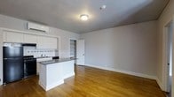 This single-floor space showcases an open-concept layout that seamlessly integrates the kitchen and living areas into one cohesive environment. Warm-toned hardwood floors run throughout, adding a sense of continuity and natural warmth to the interior. The neutral gray walls provide a clean, versatile backdrop suitable for a variety of design styles.

The kitchen is thoughtfully designed with crisp white cabinetry, a dark countertop island that doubles as a breakfast bar, and a decorative tile backsplash detail beneath the upper cabinets. A sink is positioned along the kitchen wall, and a dishwasher is neatly integrated into the cabinetry. A wall-mounted split air conditioning unit ensures efficient climate control within the space.

Adjacent to the kitchen, a set of built-in open shelves offers convenient storage or display space, flanked by a white door that leads to a separate room beyond. The living area is generously proportioned, offering ample room for furniture arrangement. Ceiling-mounted lighting provides even illumination throughout, and a smoke detector is also present overhead. The overall ambiance is clean, modern, and move-in ready.