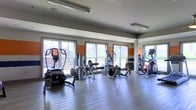 This fitness center offers a dedicated space for exercise and wellness. The room features a clean, modern aesthetic with a neutral gray and white color scheme accented by bold orange and dark horizontal stripes along the walls, adding a dynamic, energetic feel to the environment. Large windows line the perimeter, flooding the space with natural light and providing views of the surrounding urban landscape, creating an open and motivating atmosphere. The flooring consists of smooth hardwood-style planks, offering a sleek and durable surface suited for a variety of workout activities. Overhead, fluorescent lighting supplements the natural light to ensure the space remains well-lit throughout the day. The fitness center is equipped with a selection of cardio machines, including ellipticals, a stationary bike, and stair climbers, as well as strength training equipment, all arranged to allow comfortable circulation throughout the room. The space is thoughtfully laid out to maximize usability while maintaining an uncluttered feel. Overall, the fitness center presents a functional and inviting environment well-suited for daily exercise routines.