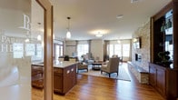 This single-floor space offers an open, airy layout that seamlessly connects a kitchenette area with expansive lounge and gathering zones. The kitchenette features rich cherry wood cabinetry topped with granite countertops and is illuminated by elegant pendant lighting, creating a warm and functional focal point as you enter the space.

The main lounge area is generously proportioned, with warm hardwood flooring extending throughout and neutral-toned walls that contribute to the inviting ambiance. Multiple seating arrangements — featuring upholstered armchairs and accent tables — are thoughtfully positioned to encourage conversation and relaxation. A striking stone fireplace anchors one end of the room, adding texture and a cozy focal point to the space.

Natural light pours in abundantly through large windows dressed with flowing neutral curtains, illuminating the room with a bright yet soft glow. French doors at the far end offer views of and access to the outdoors, further enhancing the sense of openness. Recessed and flush-mount ceiling lighting supplements the natural light, ensuring the space remains well-lit throughout the day. Overall, the design strikes a balance between elegance and comfort, making this a highly functional and welcoming communal environment.