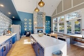 The Halstead Club clubhouse in Beverly, MA, is a thoughtfully designed amenity space that masterfully blends social gathering, recreation, and relaxation into one cohesive, light-filled environment. The architecture is immediately striking, anchored by soaring vaulted ceilings with white-trimmed gable detailing that lend an airy, cathedral-like quality to the interior. Warm-toned hardwood flooring runs throughout, unifying the open-plan layout beneath abundant natural light.

The lounge area serves as the heart of the space, centered around a dramatic floor-to-ceiling stone fireplace accented with patterned blue-and-white tilework and a rustic wood mantel. Bold teal accent walls, gold porthole-style wall décor, and a mix of plaid and leather seating give this zone a nautical-inspired character that feels both curated and inviting.

Adjacent to the lounge is a full kitchen outfitted with navy cabinetry, a generous white quartz island with bar seating, stainless steel appliances, and brass globe pendant lighting — ideal for entertaining. The game area occupies the sun-drenched windowed end of the space, where expansive floor-to-ceiling windows and French doors frame lush outdoor views of the grounds and pool. A ping pong table and shuffleboard table complete this recreational zone. The combination of natural light, distinctive design, and versatile amenities makes this clubhouse an exceptional community gathering destination.