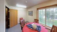 This single-floor space offers a warm and functional layout centered around a comfortable gathering room. The main area is anchored by a round dining table surrounded by wooden chairs, creating an inviting setting for conversation and casual meals. A textured ceiling with flush fluorescent lighting keeps the room well-lit, while a large multi-pane window with warm wood trim allows natural light to filter in and offers pleasant views of surrounding greenery and an outdoor deck area. Framed artwork adorns the cream-colored walls, adding a touch of character to the space.

An open pass-through connects the main gathering area to an adjoining corridor lined with extensive built-in bookshelves, filled with a wide collection of reading materials. This library hallway adds both a functional and welcoming element to the overall space, ideal for reading or quiet study. A small ledge shelf along the passage displays decorative items such as a globe and framed pictures, further enhancing the homey atmosphere.

Additional features include multiple interior wood-panel doors, carpeted flooring in select areas transitioning to smooth surfaces, and a small entryway with seating. The overall ambiance is calm, lived-in, and purposefully arranged to support both communal gathering and individual comfort.