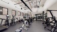 This single-floor fitness facility offers a comprehensive workout environment designed to support a wide range of exercise routines. The space features an open layout with high ceilings that expose industrial-style ductwork, piping, and structural elements overhead, lending the room a modern, utilitarian aesthetic. Bright linear pendant lighting runs throughout, ensuring the space is well-lit during all hours of use.

The flooring consists of durable dark rubber matting, ideal for high-impact activities and equipment use. Along one wall, large framed mirrors extend the visual depth of the room and assist users in monitoring their form during workouts. The walls are finished in clean white, complementing the black and grey tones of the equipment throughout.

Cardio equipment includes rowing machines, stationary bikes, and an elliptical trainer, positioned toward the front of the space. Toward the rear, a dedicated strength training zone houses a cable machine, squat rack, and a well-stocked dumbbell rack with a range of weights. The overall layout is organized and spacious, allowing for comfortable movement between stations. The facility presents a functional, modern environment suited for varied fitness goals.