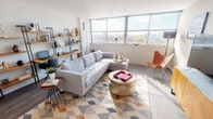 This single-floor residence showcases a thoughtfully designed open-concept layout that seamlessly connects the living, dining, and kitchen areas. The living room is bathed in natural light through large floor-to-ceiling windows, offering sweeping elevated city views. Wood-look flooring, a tufted sectional sofa, a geometric-patterned area rug, and a gold cylindrical coffee table contribute to the space's contemporary aesthetic. An open shelving unit along one wall provides both storage and display space.

The kitchen is outfitted with medium-toned wood cabinetry, white quartz countertops, a tile backsplash, and a full suite of stainless steel appliances, including a dishwasher, range, and refrigerator. A kitchen island with seating for two serves as a casual dining area, illuminated by pendant lighting overhead.

The bedroom features expansive windows that continue the cityscape views, with plush carpeting, a built-in open closet with sliding mirror panels, and ample room for furnishings. The bathroom maintains the modern design language with a wood-tone vanity, white countertop, rectangular undermount sink, and a framed mirror with track lighting above.
