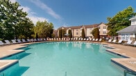 Located at Pointe of Woods in Parlin, New Jersey, this outdoor amenity area is centered around a generously sized swimming pool with crystal-clear turquoise water. The pool features gentle entry steps with handrails, making it accessible and inviting for all residents. A broad, paved sun deck surrounds the pool and is lined with an extensive row of white lounge chairs, providing ample space for relaxation and sunbathing. A lifeguard station with a shaded umbrella is positioned poolside, reflecting the community's commitment to safety and comfort. The deck also includes a dedicated seating area furnished with tables, chairs, and market umbrellas, ideal for outdoor dining and socializing. Tall, manicured arborvitae trees line the perimeter of the space, offering natural privacy screening and a lush, landscaped backdrop. A charming pavilion-style clubhouse with large arched windows and stone accents anchors one side of the area, seamlessly connecting indoor and outdoor amenities. The surrounding community buildings feature traditional architectural detailing with neutral siding and pitched rooflines. Under open skies and surrounded by mature trees, this outdoor space provides a welcoming and well-appointed recreational retreat for the Pointe of Woods community.