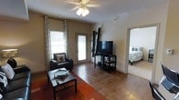 This single-story space presents a welcoming and functional layout centered around a comfortable living area. The room features warm hardwood flooring that extends throughout, lending a cohesive and inviting feel to the interior. A ceiling fan with an integrated light fixture sits centrally overhead, providing both air circulation and ambient lighting.

Natural light enters through two windows dressed with bold black-and-white patterned curtains, complementing the neutral wall tones. A glass-paneled door adds an additional source of daylight while offering access to the exterior. The living area is furnished with a dark leather accent chair paired with a low-profile coffee table and an area rug, creating a defined and cozy seating zone.

A sleek, angular media shelf unit stands against one wall, housing a flat-screen television and additional storage, adding a modern touch to the space. Through an open doorway, a bedroom is visible, featuring a bed with neutral bedding and continued hardwood flooring, suggesting a consistent design language throughout the home. Overall, the space combines practical design with a warm, lived-in ambiance.
