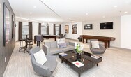 Located at 20 E 19th St in Bayonne, NJ, this single-floor amenity lounge is designed for recreation and socializing, combining modern aesthetics with a versatile layout. The space centers around a full-size billiards table set beneath a striking sputnik-style chandelier, with natural light streaming in through large windows dressed with dark curtains. Adjacent to the game area, a comfortable lounge zone features plush gray seating, a sleek black coffee table, and a wall-mounted flat-screen TV positioned across from a contemporary metallic console. A textured dark accent wall adds visual depth to the lounge area, complemented by bold, large-scale framed artwork that lends an energetic, curated feel throughout. A dedicated hallway corridor extends the entertainment offerings with a shuffleboard table, an additional wall-mounted TV, and more framed photography-style art. Recessed lighting throughout ensures a bright, welcoming atmosphere, while the neutral-toned, striped carpeting ties the various zones together cohesively. The overall design balances an upscale, modern sensibility with functional amenities, making this space well-suited for leisure and community engagement.