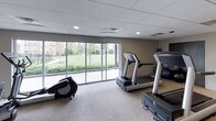 This fitness center offers a comprehensive workout environment spread across a single level, designed to accommodate a variety of exercise routines. The space features a drop-panel ceiling with recessed lighting throughout, providing consistent, even illumination across the workout areas. Neutral-toned carpet flooring and clean white walls give the room a bright, welcoming atmosphere.

The cardio area is well-appointed with multiple treadmills, elliptical trainers, and a stationary bike, several of which are positioned in front of a large wall-mounted flat-screen television — ideal for staying entertained during workouts. A standout feature of the space is the expansive floor-to-ceiling windows along one wall, which flood the room with natural light and offer open views of a lush green outdoor area, creating an energizing environment for exercise.

A dedicated strength training zone includes a dumbbell rack with a full range of free weights, a weight bench with protective floor mats, and additional open floor space for stretching or bodyweight exercises. An adjacent room, visible through a glass partition, suggests the availability of additional facilities nearby. The overall layout is thoughtfully organized, balancing equipment density with ample room to move freely.