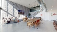 Located at 13448 Beach Ave in Los Angeles, CA 90066, this single-floor space delivers a thoughtfully designed environment that blends industrial aesthetics with modern comfort. The main common area commands attention with soaring double-height ceilings, exposed ductwork, and polished concrete floors that lend an authentic industrial character. A bold patterned feature wall anchors the lounge zone, where eclectic seating arrangements — including upholstered chairs and intimate café-style tables — invite both collaboration and casual gathering. A large communal wooden table with mixed seating serves as a central focal point, ideal for group work or social interaction. A wall-mounted multi-panel display adds a media-ready element to the space. An open staircase with glass paneling leads to a mezzanine level overlooking the main floor, adding architectural depth and visual interest. Pendant lighting with geometric cage fixtures illuminates the kitchen, which features warm wood cabinetry, dark stone countertops, a speckled tile backsplash, a professional-grade range, and a central island with bar seating — making it both functional and refined. Natural light filters throughout, enhancing the bright, airy atmosphere of this versatile Los Angeles property.