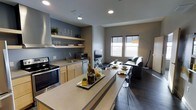 This single-floor space showcases an open-concept layout that seamlessly connects the kitchen, dining, and living areas. The kitchen is well-appointed with stainless steel appliances, including a range and microwave, complemented by light wood cabinetry, open shelving for display and storage, and a deep sink. A generous island with a smooth countertop serves as both a prep area and a casual dining spot, with bar-height seating adding functional versatility.

The adjoining dining area flows naturally into the living space, which features dark hardwood-style flooring and a neutral gray color palette throughout. The living room is anchored by a sleek dark sofa and accented with a geometric-patterned area rug that adds visual interest underfoot. A stylish arc floor lamp provides warm ambient lighting. Multiple windows allow abundant natural light to fill the space, enhancing the open and airy atmosphere.

Decorative touches such as a wall-mounted clock, framed artwork, and curated shelf displays add personality without overwhelming the clean, contemporary aesthetic. The overall ambiance is modern, cohesive, and thoughtfully designed for comfortable everyday living.