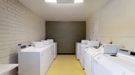 This shared laundry facility is a clean and efficiently arranged space designed to accommodate multiple users at once. The room features a symmetrical layout, with a row of full-sized top-loading washing machines and dryers lining each side wall, providing ample capacity for laundry needs. The flooring consists of durable yellow tile, offering a practical and easy-to-clean surface. The walls are finished in painted white brick, with a contrasting unpainted gray brick accent wall serving as a focal point at the far end of the room, adding an industrial character to the space. Overhead, flush-mounted fluorescent lighting ensures the room is well-lit throughout. Posted signage near the machines provides users with guidelines and instructions for proper use. The overall layout is straightforward and functional, maximizing the use of the available floor space while maintaining clear pathways between the two rows of appliances. The space is designed with practicality in mind, offering a tidy and accessible environment for laundry tasks.