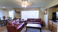This single-floor residence offers a comfortable and functional living environment designed with everyday convenience in mind. The living room serves as the heart of the space, featuring plush carpeted flooring that adds warmth underfoot. A large window dressed with horizontal blinds allows generous natural light to fill the room, creating a bright and inviting atmosphere. A ceiling fan with an integrated light fixture is centrally positioned overhead, providing both air circulation and ambient lighting. The living area is well-suited for relaxed gatherings, with enough space to accommodate a full sofa and loveseat arrangement paired with a sleek coffee table. A decorative metal shelving unit adds a stylish organizational element to the corner of the room. The textured ceiling adds a classic residential character throughout. The overall ambiance is warm and lived-in, offering a practical layout that balances comfort with functionality. The space is ideal for those seeking a straightforward, single-level home with a cozy and approachable feel throughout its interior.