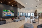 This single-floor community clubhouse is designed with a contemporary open-concept layout that maximizes both function and visual appeal. Soaring ceilings throughout the main space create an expansive, airy atmosphere, complemented by a striking combination of warm wood ceiling panels and crisp white walls. The flooring transitions seamlessly between large-format tile and light hardwood, grounding each distinct activity zone.

The lounge area anchors one end of the space, featuring a generous sectional sofa, a patterned area rug, and a large-format video display wall framed in wood veneer. Abstract artwork adorns the deep charcoal accent wall, adding a gallery-like quality to the seating nook. Adjacent to this area, a full-length shuffleboard table sits beneath floor-to-ceiling windows, flooding the zone with natural light and offering views of the surrounding outdoor landscape.

Toward the rear of the clubhouse, a pool table occupies a central position beneath a dramatic sputnik-style chandelier. A long communal bar-height table with upholstered seating runs alongside a built-in wet bar with under-counter refrigeration and wall-mounted display shelving. Oversized glass garage doors on one side of the room open to reveal outdoor pool and courtyard views, while multiple large sliding windows maintain a bright, connected feel throughout the space.