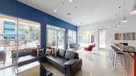 This single-floor communal space is designed with a contemporary aesthetic that blends bold color, open layout, and seamless indoor-outdoor connectivity. Striking cobalt blue accent walls contrast sharply against crisp white ceilings and walls, creating a vibrant, energetic atmosphere throughout.

The main lounge area is generously proportioned, featuring polished concrete flooring that reflects natural light flooding in through expansive floor-to-ceiling sliding glass doors. These doors open directly onto a sun-drenched outdoor courtyard equipped with patio furniture and umbrella-shaded tables, offering an effortless transition between interior and exterior spaces. Multiple seating arrangements are thoughtfully distributed across the lounge, including a large sectional leather sofa, a gray contemporary couch, and a pair of bold red accent chairs arranged around a geometric-patterned area rug.

A dedicated media nook features a wall-mounted flat-screen television recessed into the blue accent wall, flanked by a pair of sleek black egg-shaped swivel chairs and a colorful striped area rug, creating an intimate entertainment corner.

The fully equipped kitchen is a standout feature, showcasing dark cabinetry with glossy black countertops, stainless steel appliances including a range, oven, and refrigerator, and pendant lighting hanging from high ceilings. Open shelving adds a functional yet decorative element, and colorful artwork accents the walls. The space as a whole is ideal for community gathering and everyday living.