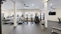 Located at 307 Karl Linn Dr in North Chesterfield, VA, this fitness center is a comprehensively outfitted facility designed to support a wide range of workout styles. The space is organized into distinct functional zones, all unified by a clean, modern aesthetic featuring white walls, durable rubber-speckle flooring, and recessed lighting complemented by ceiling fans throughout.

The main strength training area houses an extensive rack of dumbbells spanning a wide weight range, adjustable benches, a cable machine, and large wall-mounted mirrors that enhance the sense of space and allow for form monitoring during exercises. Adjacent to this zone, a dedicated functional fitness room is accessible through glass-panel doors and features TRX suspension trainers, a heavy punching bag, kettlebells, and a colorful assortment of medicine balls on a standing rack—providing a versatile environment for dynamic and cross-training workouts.

The cardio section benefits from natural light streaming through multiple large windows, offering views of the surrounding exterior. It is equipped with treadmills, elliptical machines, upright and recumbent bikes, and a stair climber. Wall-mounted flat-screen televisions are present throughout the space, enhancing the workout experience. The overall ambiance is bright, open, and well-organized.
