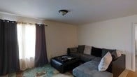 This single-floor space offers a straightforward and functional layout centered around a comfortable living area. The living room serves as the heart of the space, furnished with a generously sized gray sectional sofa that provides ample seating. A dark leather ottoman doubles as a coffee table, adding both practicality and a contrasting design element to the room. The floor is adorned with a patterned area rug in muted tones, lending a subtle decorative touch to the space. A window dressed with dark blackout curtains allows for flexible light control, letting in natural light when desired while maintaining privacy. The walls are finished in a clean, neutral white tone that keeps the space feeling open and bright. Overhead, a simple flush-mount ceiling light fixture provides ambient illumination, contributing to the room's understated and practical aesthetic. The overall ambiance of the space is calm and modest, offering a welcoming environment suitable for everyday relaxation and living.