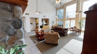 This single-story space is defined by its dramatic vaulted ceilings, which lend an airy, expansive quality to the interior. The open-concept design connects the main living area to the kitchen, creating a cohesive and functional layout. The living area is generously sized, anchored by a stone fireplace on one side and furnished with multiple seating arrangements, including patterned armchairs and a dark upholstered sofa, centered around a wooden coffee table atop a neutral area rug. The flooring transitions from stone tile near the entryway to carpet throughout the main gathering space.

Large windows along the far wall allow abundant natural light to fill the room, offering views of the surrounding greenery outdoors. Two ceiling fans with light fixtures hang at varying heights, serving both the lower and upper portions of the vaulted space. The kitchen, visible through an arched pass-through opening, features wooden cabinetry, a refrigerator, and counter seating, maintaining visual connectivity with the living area. A dining nook with chairs is positioned near the windowed wall, completing the open, communal character of the space.