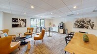 Located at 700 Woodrow St in Columbia, South Carolina, this single-floor commercial space combines contemporary design with a welcoming, professional atmosphere. The layout is thoughtfully arranged to accommodate both relaxed seating and functional gathering areas throughout.

The lounge areas are furnished with tufted caramel leather sofas and accent chairs in warm amber tones, paired with sculptural side tables and soft-hued throw pillows that create an inviting yet polished environment. Patterned carpet tiles in earthy tones run throughout, adding texture and visual interest underfoot. Recessed lighting set into drop-tile ceilings provides consistent, comfortable illumination across the space.

A wall-mounted flat-screen television anchors one of the lounge zones, while floor lamps add layered warmth. The gathering area features a beverage station equipped with a professional coffee machine, complemented by nearby high-top tables and bar-height seating — ideal for casual meetings or breaks. Large windows in this area look out onto lush greenery, bringing in natural light and a connection to the outdoors.

Private rooms with glass-paneled doors offer quieter settings accessible from the main lounge, while narrow vertical windows allow filtered light into transitional corridors. Curated wall art and floral accents complete the refined aesthetic, making this a versatile and well-appointed professional environment.