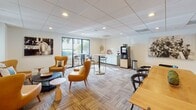 Located at 700 Woodrow St in Columbia, South Carolina, this single-floor commercial space combines contemporary design with a welcoming, professional atmosphere. The layout is thoughtfully arranged to accommodate both relaxed seating and functional gathering areas throughout.

The lounge areas are furnished with tufted caramel leather sofas and accent chairs in warm amber tones, paired with sculptural side tables and soft-hued throw pillows that create an inviting yet polished environment. Patterned carpet tiles in earthy tones run throughout, adding texture and visual interest underfoot. Recessed lighting set into drop-tile ceilings provides consistent, comfortable illumination across the space.

A wall-mounted flat-screen television anchors one of the lounge zones, while floor lamps add layered warmth. The gathering area features a beverage station equipped with a professional coffee machine, complemented by nearby high-top tables and bar-height seating — ideal for casual meetings or breaks. Large windows in this area look out onto lush greenery, bringing in natural light and a connection to the outdoors.

Private rooms with glass-paneled doors offer quieter settings accessible from the main lounge, while narrow vertical windows allow filtered light into transitional corridors. Curated wall art and floral accents complete the refined aesthetic, making this a versatile and well-appointed professional environment.