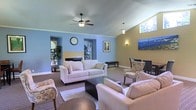 This single-story space features a generously sized living area defined by soaring vaulted ceilings that lend a sense of openness and airiness throughout. The interior design combines soft blue and warm golden-yellow wall tones, creating a visually engaging and inviting atmosphere. A ceiling fan with a light fixture is centrally mounted, complementing the overall layout while aiding air circulation.

Natural light filters into the room through windows, including a strategically placed upper window that draws light into the vaulted ceiling area. Recessed wall niches add architectural character to the blue-painted walls, with one niche housing a decorative wall clock and another framing a potted plant.

The living area is furnished with a neutral-toned sofa, coordinating armchairs, and a dark rectangular coffee table, all arranged atop a patterned area rug. Wall sconces and pendant lighting provide warm supplemental illumination. A framed artwork and a vibrant canvas painting add decorative interest to the walls.

A dining or seating area is visible toward the rear, furnished with additional chairs and a table, further highlighting the versatile and functional layout of this comfortable space.