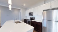 Located at 6770 Printers Blvd in New Albany, Ohio, this single-floor residence offers a thoughtfully designed living environment with modern finishes and a clean, contemporary aesthetic. The kitchen serves as a standout feature of the space, showcasing a striking two-tone cabinetry design with rich dark espresso lower cabinets contrasted against crisp white upper cabinets. White subway tile backsplash adds a classic yet modern touch to the cooking area. A full suite of stainless steel appliances — including a French door refrigerator, electric range, and over-the-range microwave — provides both style and functionality. The kitchen also features a large island with a white quartz countertop and an undermount sink with a sleek arc-neck faucet, offering ample workspace and an inviting gathering spot. Recessed lighting illuminates the space efficiently, while light-toned walls and wood-look flooring contribute to a bright and welcoming atmosphere throughout. The open layout connects the kitchen seamlessly to the rest of the residence, creating a fluid and functional flow. Overall, the space combines practical design with modern sensibilities, making it well-suited for everyday living.
