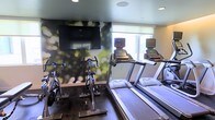 This fitness center offers a well-appointed workout environment designed to support a range of cardio-focused exercise routines. The room features multiple treadmills equipped with individual digital consoles, allowing users to track their workout metrics with ease. Alongside the treadmills, stationary cycling bikes provide an additional cardio option, making the space versatile for different training preferences. A large flat-screen television is wall-mounted prominently, offering entertainment during workouts. The room is enhanced by a striking nature-inspired mural wallpaper that creates a vibrant and energizing backdrop, depicting soft bokeh greenery that adds depth and visual interest to the space. A window allows natural light to filter in, brightening the room and contributing to an open, welcoming atmosphere. Recessed ceiling lighting supplements the natural light, ensuring the space remains well-lit at all times. The dark flooring provides a clean, modern contrast to the lighter walls and equipment. Overall, the fitness center is thoughtfully designed to create a motivating environment that balances functionality with aesthetic appeal, making it an inviting space for daily exercise.