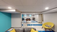 This single-floor space is designed as a dynamic recreational and social environment, combining multiple activity zones within an open, well-lit layout. The flooring throughout the main area features a striking white-and-blue striped vinyl tile pattern that anchors the space with a bold, energetic aesthetic.

The primary lounge area is furnished with a grey sofa, a patterned area rug, and mid-century-style yellow accent chairs, set against a vibrant teal feature wall adorned with a cityscape artwork. Recessed lighting in a drop-panel ceiling provides consistent, even illumination throughout.

Adjacent to the lounge, the game zone houses a full-size pool table and a foosball table, offering ample room for play. Small hopper windows along the upper walls allow modest natural light into the space.

A separate nook with a teal accent wall, a round lounge chair, and a blue area rug provides a quieter retreat. An additional media area includes a wall-mounted flat-screen television above a work table, with a private room accessible through a nearby door. Together, these zones create a versatile, modern space well-suited for both relaxation and entertainment.
