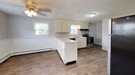 This two-floor space is finished in a neutral gray palette that lends a clean, understated character throughout. The main level features wood-look flooring that flows through the living area and into the kitchen. The living space is bright and open, with large windows admitting generous natural light and views of the surrounding neighborhood. A staircase connects the two floors, and a wall-mounted air conditioning unit provides supplemental cooling on this level.

The kitchen is well-appointed with white cabinetry, light countertops, a double stainless steel sink, and a stainless electric range. A peninsula provides additional counter space and a natural division between the kitchen and adjacent areas. A ceiling fan with a light fixture adds both function and airflow to this space.

The upper level hosts multiple carpeted bedrooms, each finished with the same soft gray walls and outfitted with baseboard heating. Bedrooms benefit from multiple windows, allowing ample natural light. One bedroom includes a wall-mounted air conditioning unit for independent climate control. Closet space is provided with full-width reach-in closets featuring hanging rods.

The bathroom includes a tub-shower combination, a window for natural light and ventilation, and consistent wood-look flooring tying it to the rest of the home.