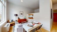This single-floor unit offers a well-organized layout that maximizes functionality through clean, modern design. Light-toned hardwood flooring flows throughout the main living spaces, lending a cohesive and airy feel. The open-plan living and dining area is generously lit by large windows that frame views of the surrounding streetscape, filling the space with abundant natural light. A white dining table paired with wire-frame chairs anchors the dining zone, while the living area features mid-century modern furnishings, including a warm wood credenza and curated wall art that add character and depth. The galley-style kitchen runs along one side of the unit, outfitted with white cabinetry, white countertops, stainless steel appliances including a refrigerator and range, and a single-basin sink — providing an efficient and stylish cooking space. A hallway connects the main living area to the private quarters, where a well-appointed bedroom showcases a striking reclaimed wood headboard with a chevron pattern, creating a warm and inviting focal point. The bathroom features hexagonal tile flooring, a wood-toned vanity with a white countertop, and a large mirror, while a stacked washer and dryer unit provides the added convenience of in-unit laundry. Throughout, the palette of crisp whites and neutral tones creates a calm, contemporary ambiance.