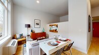 This single-floor unit offers a well-organized layout that maximizes functionality through clean, modern design. Light-toned hardwood flooring flows throughout the main living spaces, lending a cohesive and airy feel. The open-plan living and dining area is generously lit by large windows that frame views of the surrounding streetscape, filling the space with abundant natural light. A white dining table paired with wire-frame chairs anchors the dining zone, while the living area features mid-century modern furnishings, including a warm wood credenza and curated wall art that add character and depth. The galley-style kitchen runs along one side of the unit, outfitted with white cabinetry, white countertops, stainless steel appliances including a refrigerator and range, and a single-basin sink — providing an efficient and stylish cooking space. A hallway connects the main living area to the private quarters, where a well-appointed bedroom showcases a striking reclaimed wood headboard with a chevron pattern, creating a warm and inviting focal point. The bathroom features hexagonal tile flooring, a wood-toned vanity with a white countertop, and a large mirror, while a stacked washer and dryer unit provides the added convenience of in-unit laundry. Throughout, the palette of crisp whites and neutral tones creates a calm, contemporary ambiance.