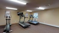 This single-floor fitness space is designed to support an active lifestyle with a clean, functional layout. The room is equipped with two commercial-grade treadmills positioned to make the most of the available floor area, offering ample space for users to exercise comfortably without feeling crowded. A wall-mounted flat-screen television is installed at an optimal viewing height, allowing users to stay entertained during their workouts. The room is finished with neutral beige walls and carpeted flooring that provides cushioning underfoot. Bright fluorescent overhead lighting ensures the space is well-illuminated throughout. A window along one wall allows for a degree of natural light and ventilation, complemented by a built-in climate control unit beneath it, ensuring year-round comfort. Baseboard heating runs along the perimeter, adding to the room's climate management capabilities. A fitness reference poster is displayed on the wall, offering workout guidance. Overall, the space is straightforward, practical, and well-appointed for cardiovascular training.