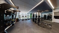 This contemporary fitness center is thoughtfully designed to support a full range of workout routines within a single, open-plan floor. The space is finished with large-format dark tile flooring throughout, providing a sleek, durable surface that anchors the room's modern aesthetic. Linear LED lighting runs along the ceiling, complementing the natural light that floods in through expansive floor-to-ceiling windows, which offer views of the surrounding urban streetscape and neighboring buildings.

The main workout area is generously proportioned, housing a dedicated cardio zone equipped with treadmills, stationary cycling bikes, and stair climbers. A strength training section features a multi-functional cable machine, a squat rack with free weights, and a well-stocked dumbbell rack accompanied by a weight bench. Additional functional training equipment includes a rowing machine and TRX suspension trainers mounted to a dedicated rack. Medicine balls and stability balls are also available, rounding out the functional fitness offerings.

A bold artistic mural in dark, abstract tones adorns one wall, while vibrant green accent walls and a striking geometric patterned door panel add visual energy to the interior. Full-length mirrors are strategically placed to enhance spatial depth and support form monitoring. A built-in cabinetry unit with countertop and storage completes the space, offering a convenient station for users.