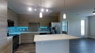 This single-floor space showcases a contemporary kitchen design that serves as the focal point of the layout. The kitchen is appointed with flat-front cabinetry in a warm wood-tone finish, providing ample upper and lower storage. A mosaic tile backsplash in muted blue and grey tones adds texture and visual interest along the full length of the cooking wall, complemented by subtle teal LED under-cabinet lighting that creates a striking ambient glow. Stainless steel appliances, including a French door refrigerator with an in-door water and ice dispenser and a built-in range, lend a polished, modern aesthetic. The spacious white kitchen island anchors the space, offering additional prep surface and a natural gathering point. Overhead, a combination of track lighting and cylindrical pendant lights provides layered illumination throughout the kitchen. Dark hardwood-style flooring grounds the space and contrasts cleanly with the bright white island countertop. A window near the adjacent area allows natural light to filter in, enhancing the open, airy feel of the overall layout.