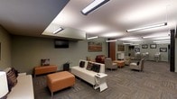 This single-floor lounge space is thoughtfully designed to offer a comfortable and inviting environment. The layout is anchored by a curated arrangement of contemporary furniture, including a cream-colored sofa adorned with decorative accent pillows, complemented by rich burnt-orange ottomans that add warmth and a pop of color throughout the room. A red leather accent chair adds a bold contrast, while additional seating areas are visible in the background, suggesting the space accommodates a variety of group sizes. The flooring features a neutral, patterned carpet that grounds the room with a subtle texture. Above, a striking suspended ceiling design frames rows of modern linear pendant lights, casting bright, even illumination throughout. The walls are painted in a muted sage green tone, providing a calm backdrop for the wall-mounted flat-screen television and framed artwork pieces that add visual interest. A mirrored wall element at the rear of the space enhances the sense of depth and openness. The overall ambiance is clean, modern, and functional, making this lounge well-suited for casual gatherings, relaxation, or community use.